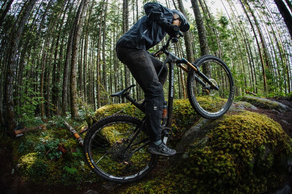 Mountain biker balancing on mossy rocks in dense forest wearing black gear and helmet.