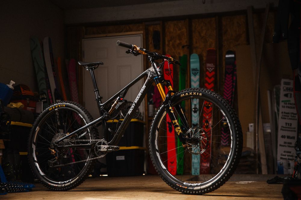 A high-end full-suspension mountain bike displayed in a dimly lit garage workshop with skis in the background.
