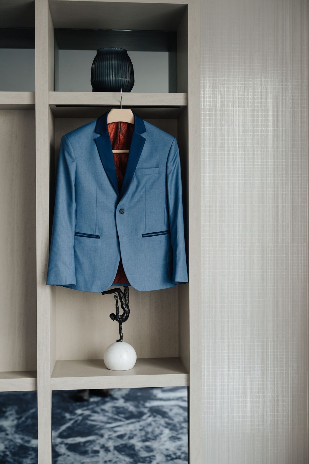 A blue suit jacket hanging on a wooden hanger inside a modern wardrobe with shelves and decorative items.
