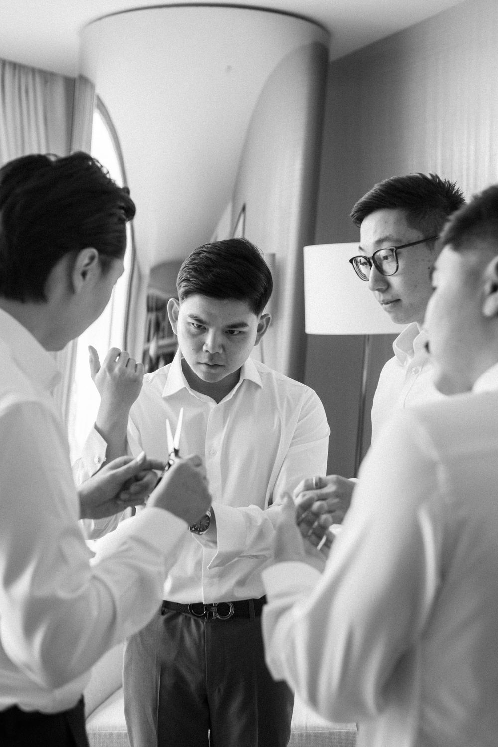 Black and white photo of groomsmen helping each other prepare in dress shirts before a wedding ceremony.