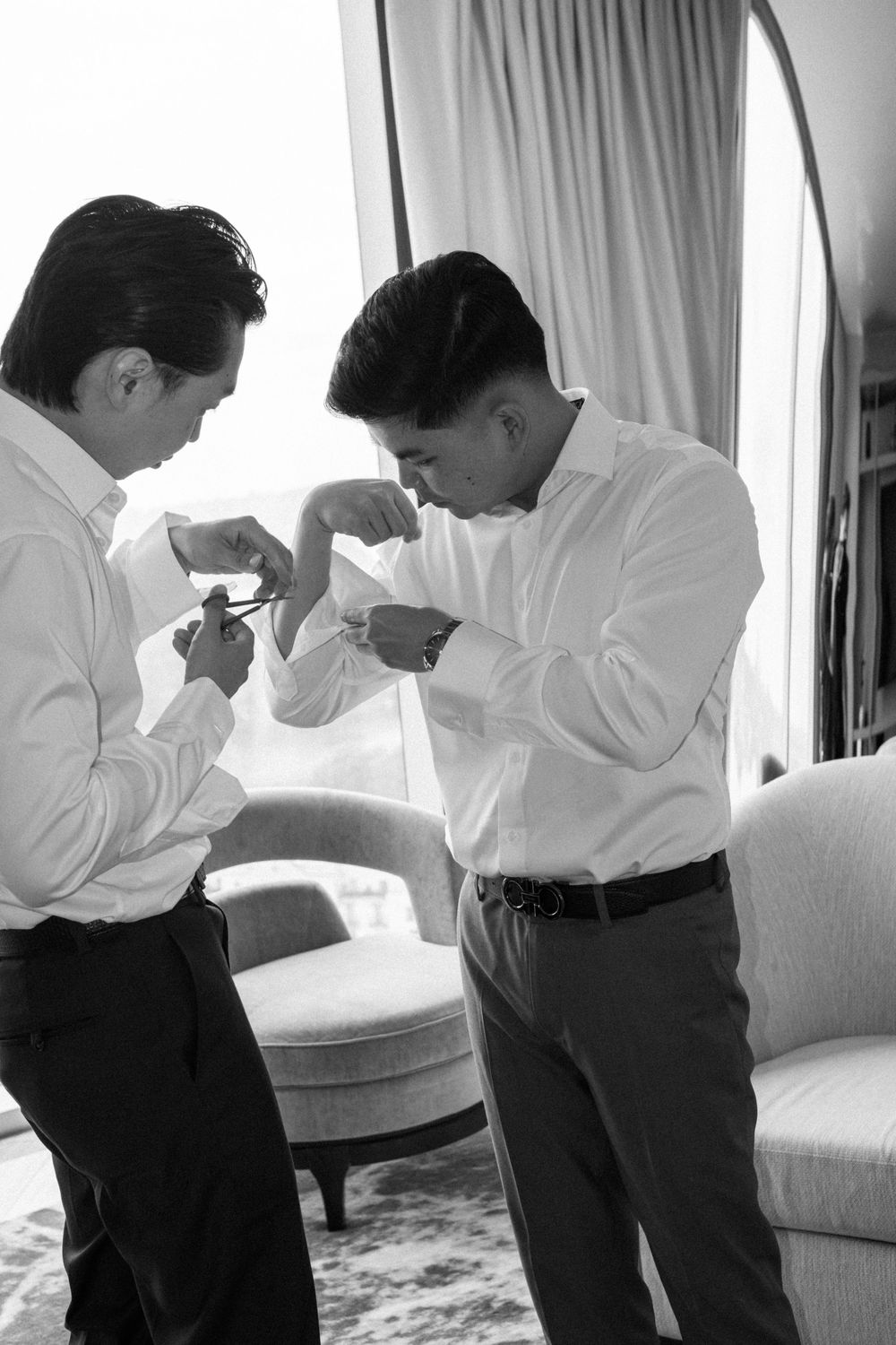 Two men in white dress shirts helping each other with cufflinks in an elegant room, black and white photo.