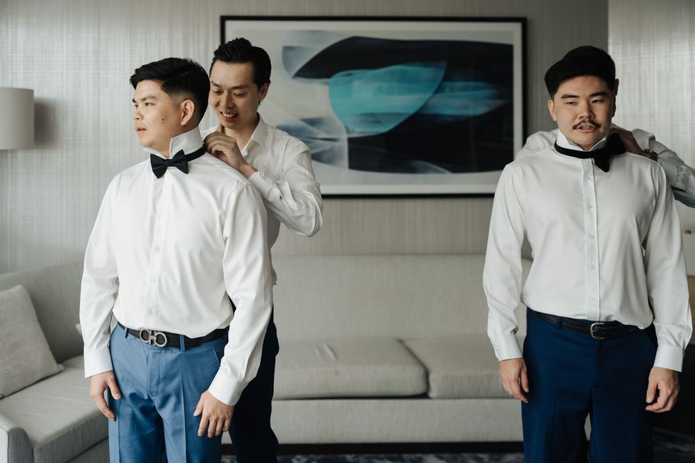 Three men in white dress shirts and bow ties prepare for a formal event in an elegant hotel room.