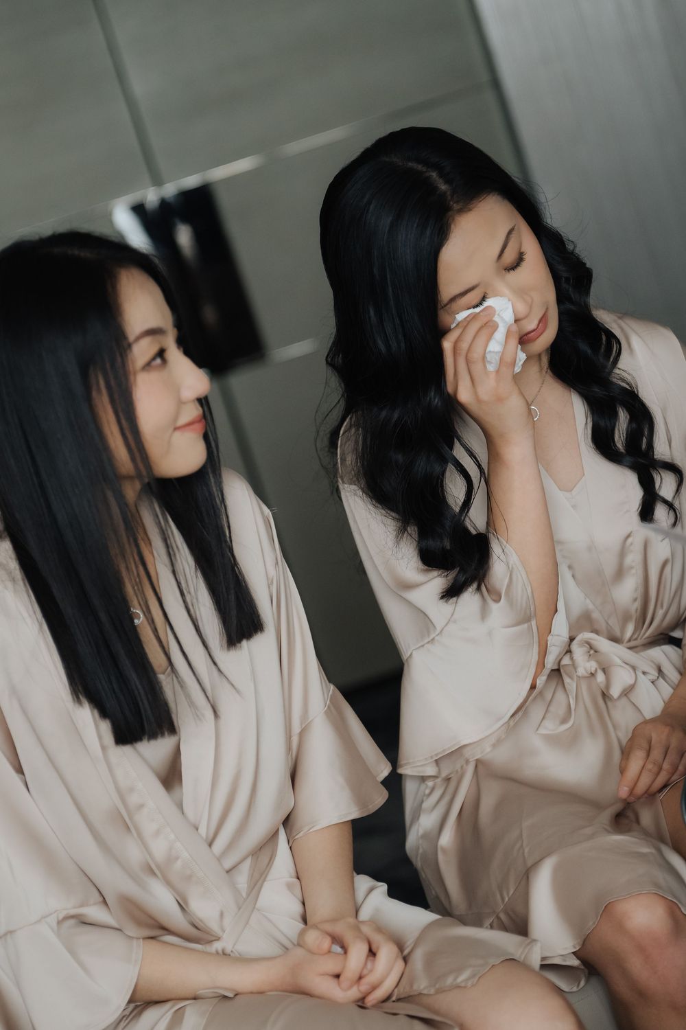 Two women in satin robes share an emotional moment, one wiping tears with a tissue during bridal preparations.