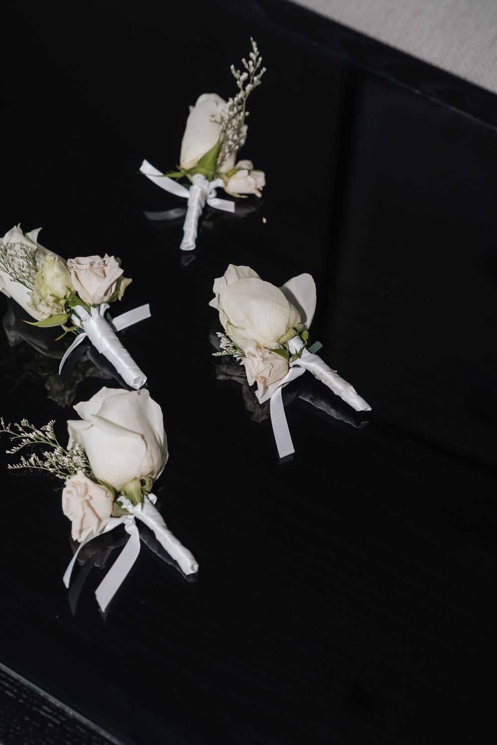 Four white rose boutonnieres with ribbon ties arranged on a dark black surface for a wedding.