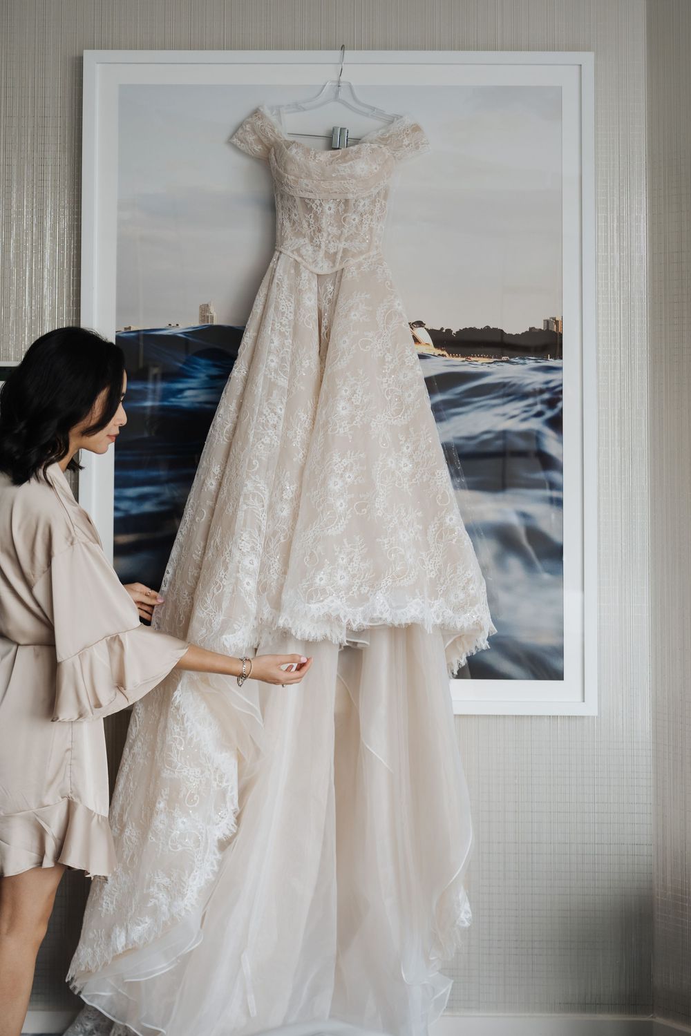 Bride admiring elegant lace off-shoulder wedding gown hanging on a frame in a bright hotel room.