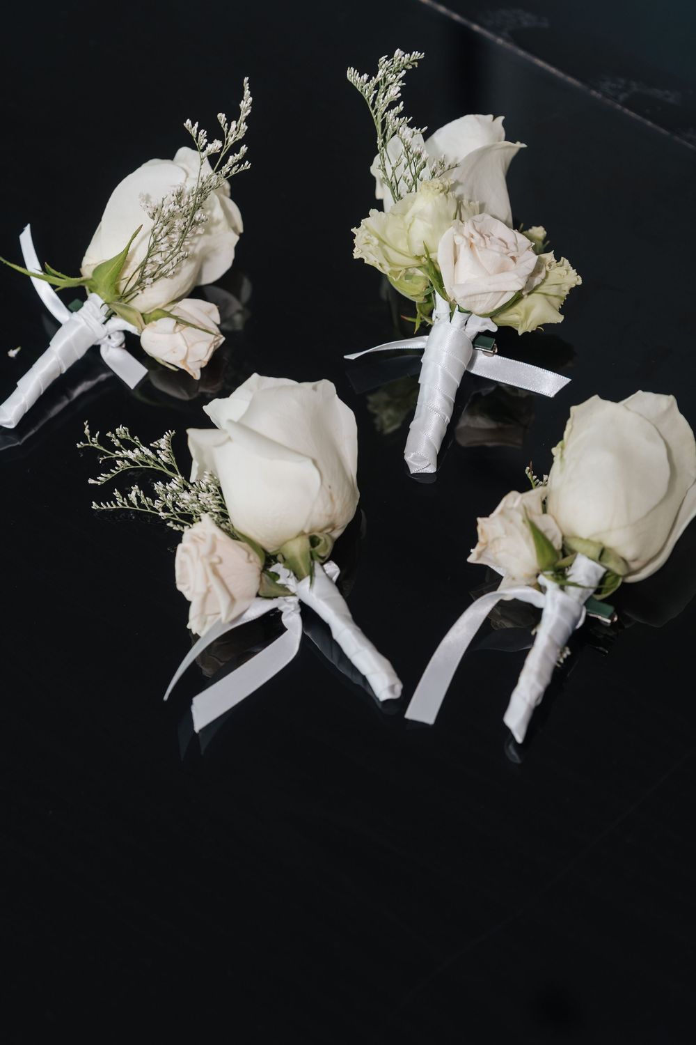 Four white rose boutonnieres with ribbon ties arranged on a dark reflective surface for a wedding.