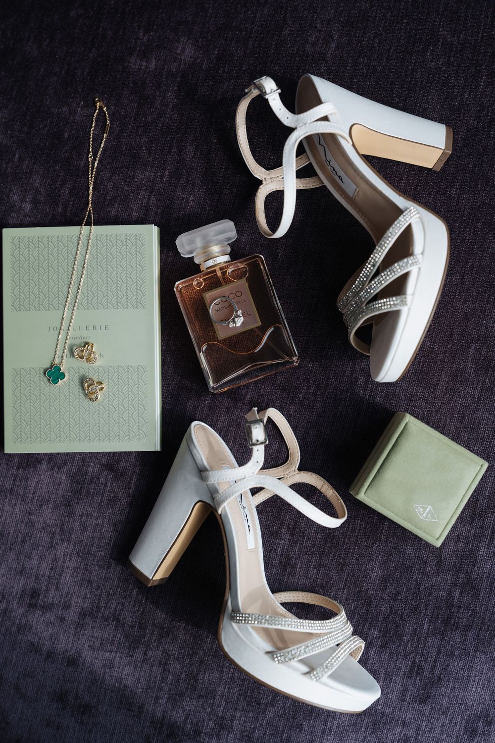White bridal heels with crystal straps, Chanel perfume, gold necklace, and green accessories on dark velvet.