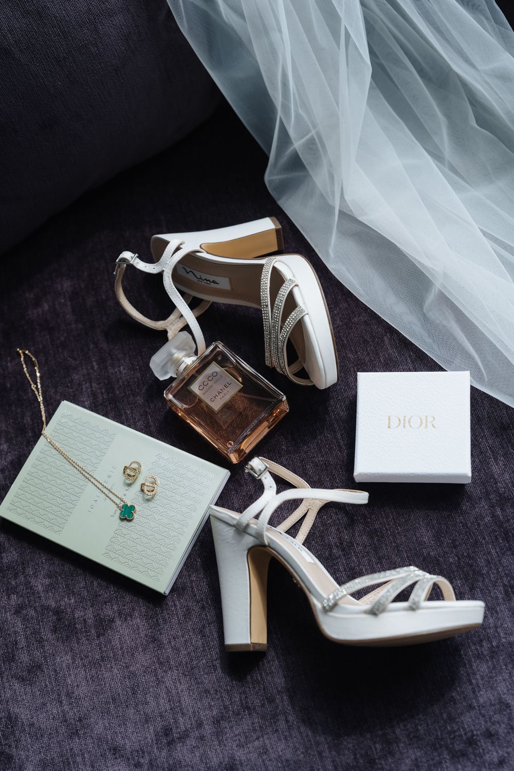 White bridal heels with Dior box, gold jewelry, perfume bottle, and tulle veil on gray velvet surface.