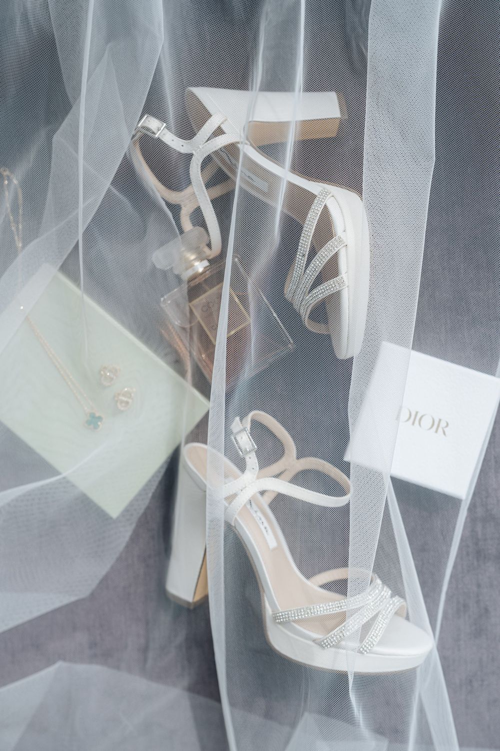 White bridal heels with crystal straps on tulle veil beside Dior box and pearl jewelry for wedding day.