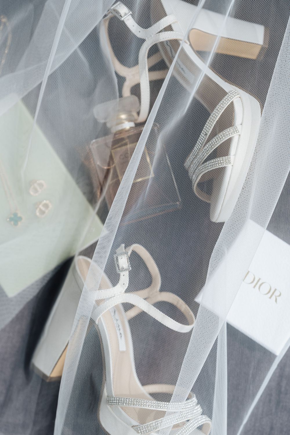 Elegant white bridal heels with crystal straps beside a Dior box, perfume bottle, and jewelry under a sheer veil.