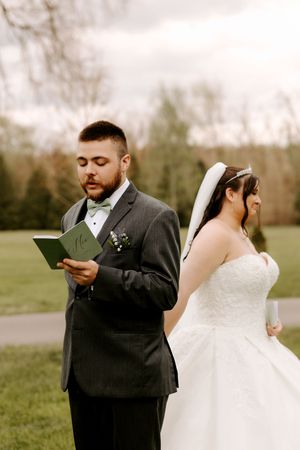 Groom in gray suit reads letter while bride in white gown stands back-to-back with him outdoors.