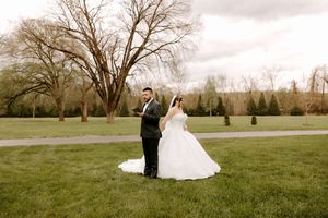 Bride and groom standing together on lush green lawn with large trees and manicured hedges in background.
