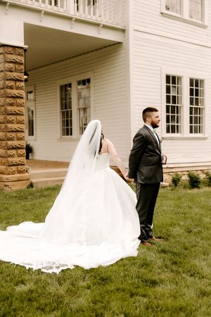 Bride in flowing white gown and veil holds groom's hand in dark suit outside elegant white venue on green lawn.