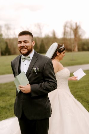 Smiling groom in dark suit holds letter while bride in white gown reads hers back-to-back outdoors.