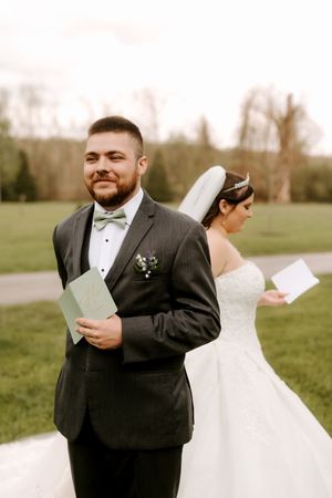 Groom in gray tuxedo smiles holding vows card while bride in white gown stands back-to-back with him outdoors.