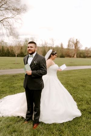 Bride and groom stand back-to-back on green lawn reading letters to each other before wedding ceremony.