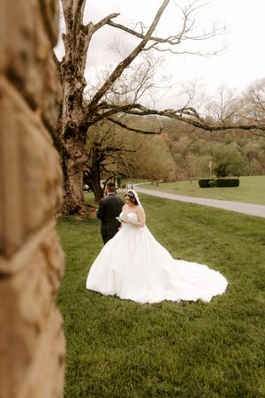 Bride in white ballgown and groom in dark suit share a romantic moment on lush green lawn near a large tree.