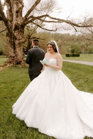 Bride in strapless ball gown reads vows outdoors as groom stands with back turned near large tree.