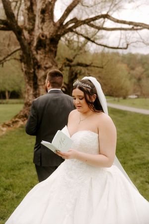 Bride in strapless lace ball gown reads vow letter while groom stands with back turned near large tree.