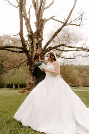 Bride in a white ballgown reads vows outdoors near a large tree while groom stands with his back turned.