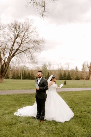 Bride and groom stand back-to-back reading vow letters in a lush green park on their wedding day.