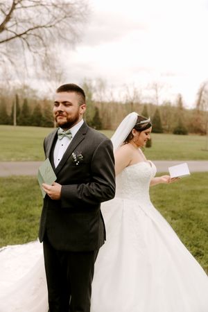 Bride and groom stand back-to-back reading vows outdoors, groom in gray tuxedo, bride in white ballgown with veil.