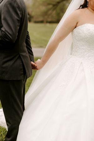 Close-up of bride in strapless lace gown holding hands with groom in dark suit outdoors.
