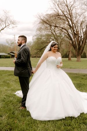Bride in white ballgown holds grooms hand outdoors on green grass with bare trees in background.