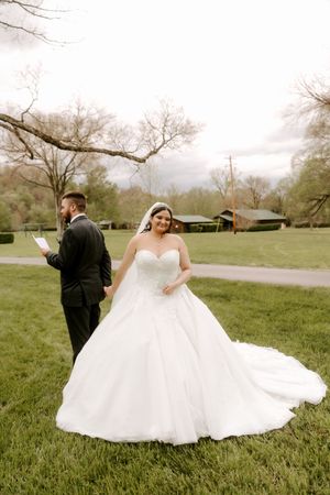 Bride in white ball gown smiles at camera while groom reads vows outdoors on green lawn.