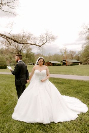 Bride in strapless white ball gown with cathedral veil stands on lawn while groom reads letter behind her.