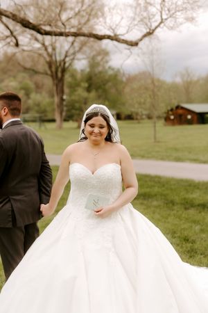 Smiling bride in strapless lace ball gown walks with groom in park on wedding day.