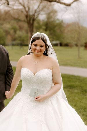Smiling bride in strapless lace ball gown holds vow booklet outdoors on wedding day.