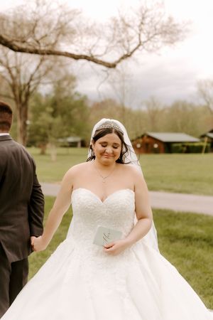Emotional bride in strapless lace ballgown and crystal tiara reads wedding vows outdoors on green lawn.