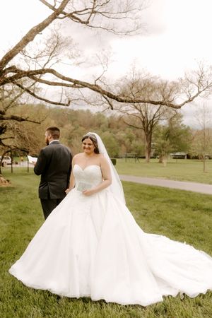 Smiling bride in strapless ball gown with long veil stands before groom in black suit outdoors.