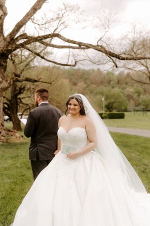 Bride in strapless ball gown and veil smiles outdoors while groom stands with his back turned beneath a tree.