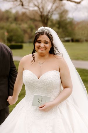 Bride in a strapless lace ballgown with veil and pearl headpiece holds vow booklet during first look ceremony.