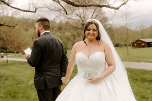 Smiling bride in strapless white ballgown and veil holds grooms hand during outdoor wedding ceremony.