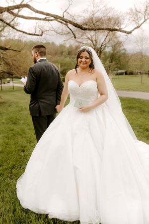 Bride in white ballgown with veil smiles outdoors while groom in black suit stands with back turned on green lawn.