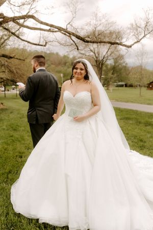 Radiant bride in white ball gown and veil smiles as groom reads vows in green park.