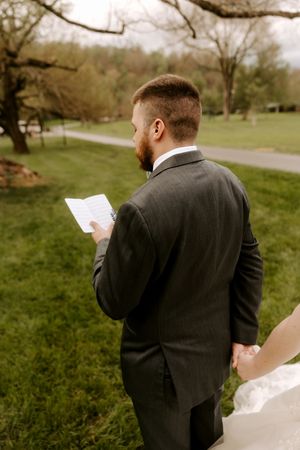 Groom in dark suit reading wedding vows from paper while holding brides hand outdoors.