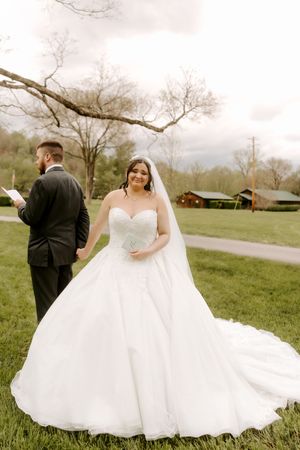 Smiling bride in a strapless white ballgown with cathedral veil stands beside groom reading vows outdoors.
