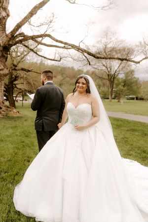 Bride in strapless lace ball gown and veil poses with tuxedo groom under bare tree.
