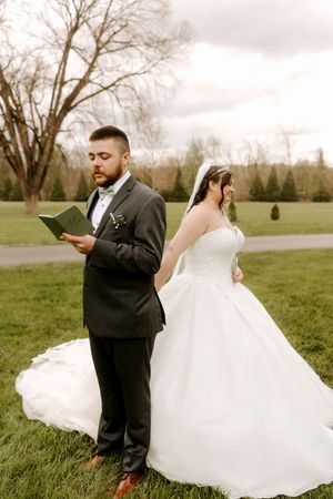 A groom in a dark suit reads vows while bride in white gown stands back-to-back with him outdoors.