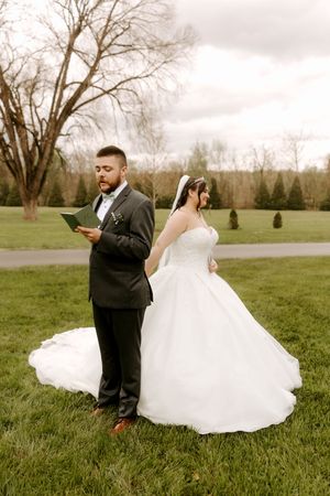 Bride and groom stand back-to-back reading private vows on green lawn before ceremony.