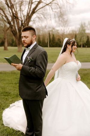 Bride and groom stand back-to-back reading private vows on green grass, bride smiling in white ballgown.