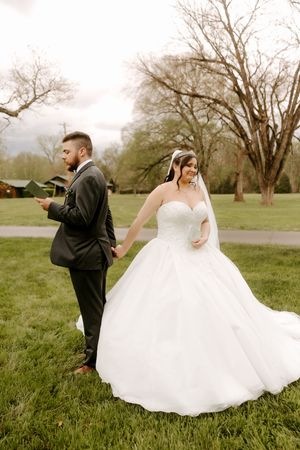 Groom in dark suit holds bride's hand as she smiles in a flowing white ballgown with veil outdoors.