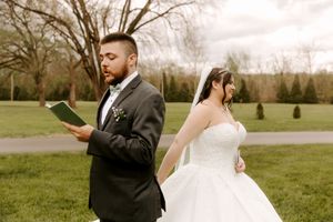 Bride in white strapless gown and groom in black suit reading vows back-to-back outdoors on green lawn.