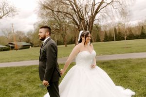 Bride in white ball gown and groom in dark suit hold hands walking across green lawn.