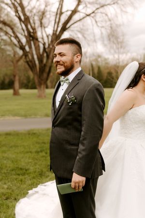Smiling groom in gray suit with green bow tie and boutonniere stands beside bride in white gown outdoors.