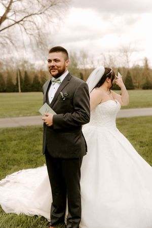 Groom in gray tuxedo holds vows while bride in white ballgown and veil stands behind him during outdoor first look.